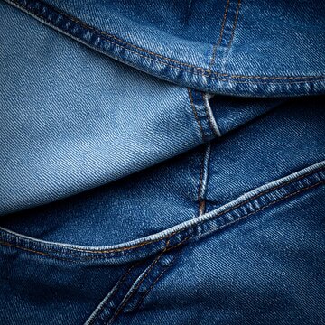 Close-up of denim fabric, showing seams and textures. Different shades of blue with contrasting stitching, folds and layers creating a visual pattern