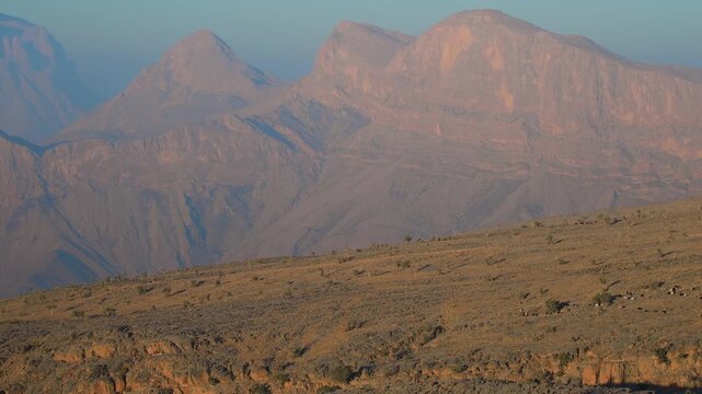 A wide-angle shot captures the rugged, arid beauty of the Hajar Mountains in Wadi Bani Khalid, Oman, bathed in the warm, golden light of the setting or rising sun.