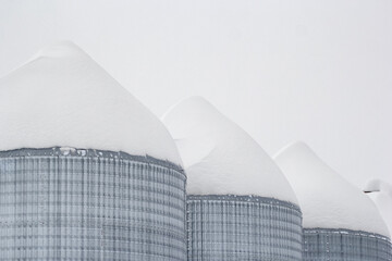Agricultural steel grain bins storing seeds in cold frosty winter. Snowbanks in the farm and on granaries after snowfall. Minimal monochrome photo.