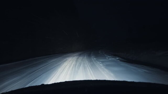 Headlights from incoming cars illuminate fresh tire tracks on rural snowcovered road with limited visibility