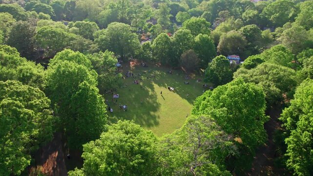 A circling aerial flying around a lush, green tree canopy surrounding an urban park as people throw the frisbee and picnic during golden hour on a bright summer day in Atlanta, Georgia.