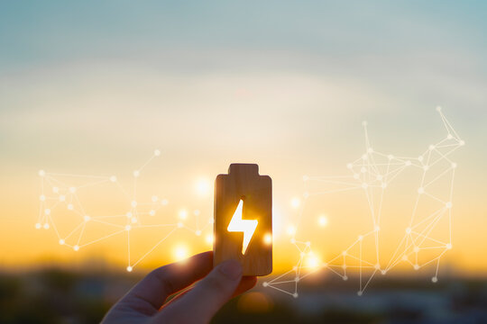 Renewable energy concept with hand holding wooden battery icon glowing at sunset. Symbol of clean energy, power storage, sustainability, future technology, green innovation, and energy management.