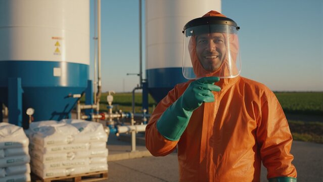 Smiling worker in orange hazmat suit pointing at camera. Professional technician in protective clothing at industrial chemical or agricultural facility