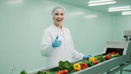 Happy food factory worker giving thumbs up for quality control. Professional woman inspecting fresh vegetables on conveyor belt in processing plant
