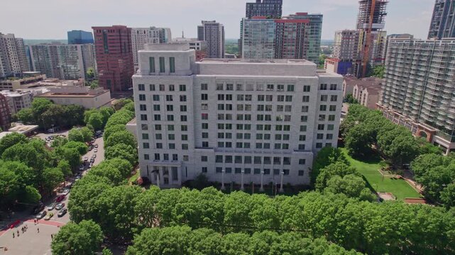Aerial approaching the granite and marble Federal Reserve building in midtown Atlanta, surrounded by a lush tree canopy with the modern city skyline behind on a bright summer day.