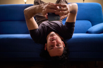 Chilled man on sofa, Casual individual reclining on sofa engrossed with mobile device, Unwinding man sits comfortably on sofa focused on his smartphone amid cozy modern decor