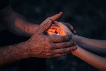 Rough scarred hands holding smooth young hands around a warm glowing light in darkness