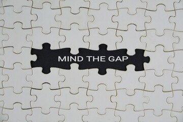 A black puzzle piece stands out amid white pieces, with the words Mind The Gap. It conveys focus on gaps, problem solving, and teamwork toward solutions.