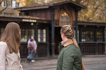 Fototapeta premium Two women stand near historic wooden tram stop in Moscow, surrounded by autumn trees, warm yellow leaves, classic urban architecture