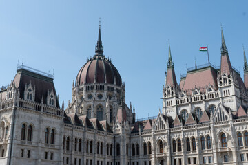 Obraz premium Hungarian Parliament Building with its central dome, neo-Gothic facade, spires, and national flag visible on the roof under a clear blue sky