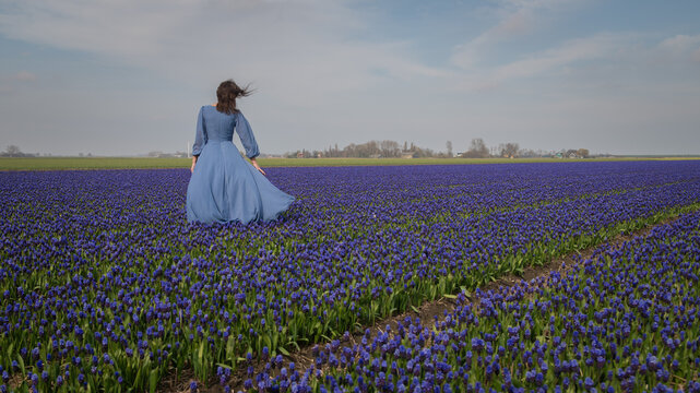 woman in blue dress standing in field of blue grape or hyacinth flowers, seen from behind under sky with clouds 