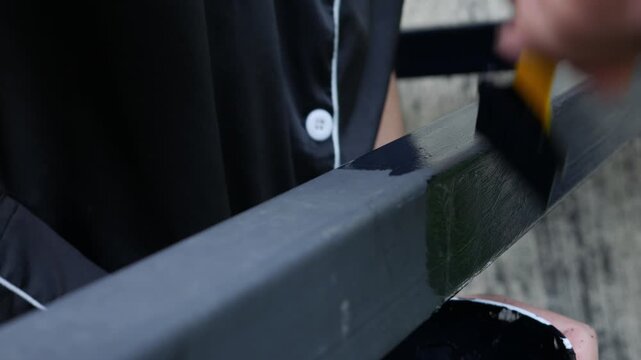Close up shot of a person's hand painting a black anti-rust protective primer or paint onto a metal frame using a small brush, home repair and maintenance concept.