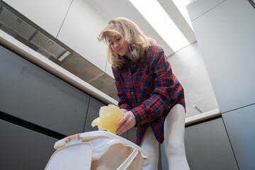 A blonde woman throws trash into a trash can in a neat modern kitchen while cleaning up after cooking. The pose is casual, demonstrating everyday household chores.