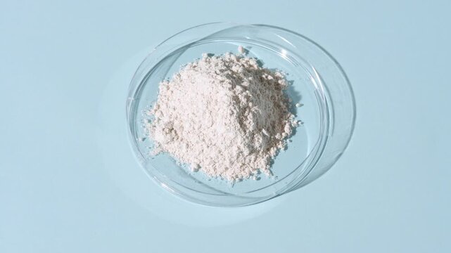 Vertical tracking shot of a stack of white fine powder inside a plastic petri dish on a pale blue background lit with orbiting light casting shifting shadows | Zinc stearate cosmetic ingredient
