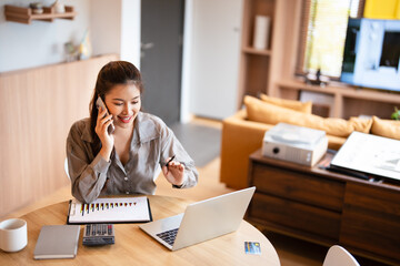 Smiling female entrepreneur discussing over mobile phone while at home office .Female business manager sharing ideas over phone call at home while looking at laptop computer screen.
