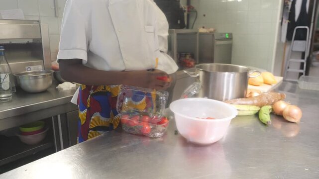 Focused action shot depicts female african chef sorting of cherry tomatoes amidst a backdrop of traditional staples like yuca and plantains as part of a kitchen montage.