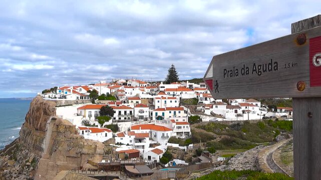 Cinematic move from the viewpoint showing the Azenhas do Mar cliffside village with red roofs, Praia da Aguda sign in the foreground, and Atlantic shore below on a cloudy day.