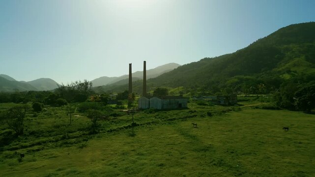 Drone flies forward toward the abandoned Santa Luiza sugar and ethanol mill in Sampaio Correia, Saquarema, Brazil. Green fields with grazing horses surround the industrial ruins; hills and mountains