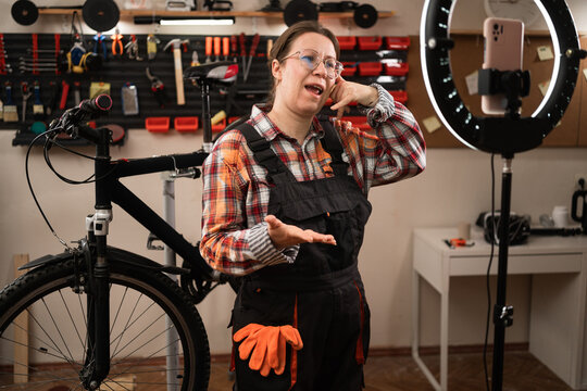Bike mechanic in vehicle maintenance job. Female worker blogger reviewing and recording video on smartphone showing call me gesture while repairing bike in bicycle workshop. - Powered by Adobe