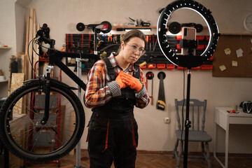 Bike mechanic in vehicle maintenance job. Female worker blogger reviewing and recording video on smartphone expressing joy and gratitude repairing bike in bicycle workshop.