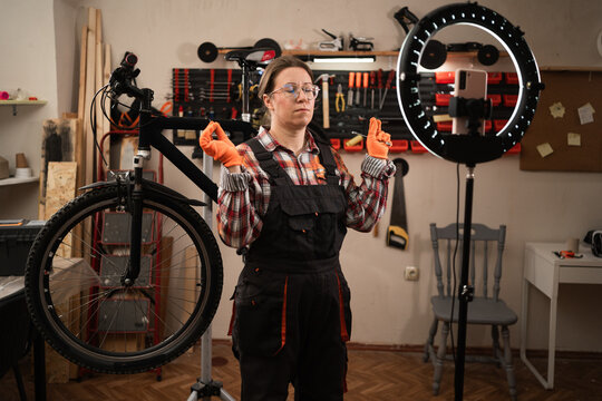 Bike mechanic in vehicle maintenance job. Female worker blogger reviewing and recording video on smartphone meditating in yoga pose repairing bike in bicycle workshop. - Powered by Adobe