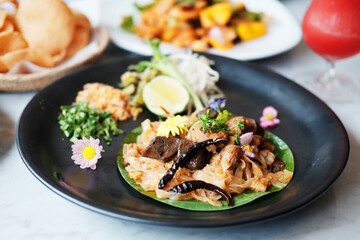 Stir fried rice noodle on banana leaf in black dish with Thai food on table