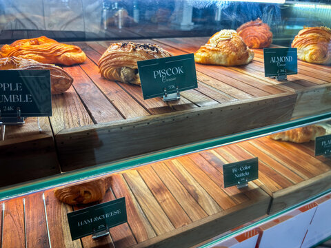 Close-up of a wooden bakery display case featuring various labeled croissants and pastries including Piscok, Almond, Ham & Cheese, and Bi-Color varieties with elegant dark green price tags.