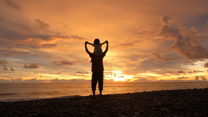 Silhouette of a father carrying his child on shoulders on the beach watching a dramatic golden sunset and cloudy sky