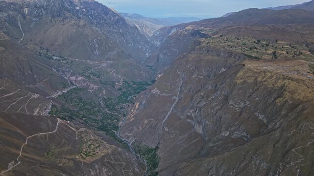 High-altitude drone of Colca Canyon Peru where narrow river opens into a vast chasm. The Rio Colca oasis, traditional farmland, rugged terrain, towering mountains, and Cabanaconde rim are visible