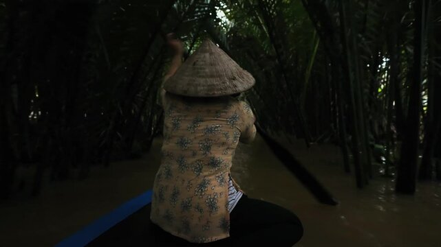 Woman in conical hat rows boat through dark, narrow canal lined with dense tropical palms, In slow motion.