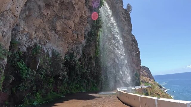 POV footage from a moving vehicle driving through Cascata dos Anjos in Madeira, Portugal, showing a coastal road where a waterfall flows directly across the roadway beside the Atlantic Ocean