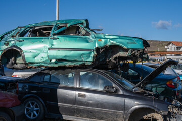 a crushed teal-green station wagon stacked precariously on top of a wrecked black sedan in a crowded salvage yard under a blue sky.