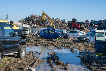 a muddy salvage yard featuring deep, water-filled tire ruts in the foreground, leading towards several wrecked vehicles and a massive scrap pile worked by a distant yellow crane.