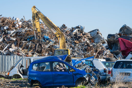 a yellow excavator crane with a grapple claw sorting through a massive mountain of scrap metal, looming over a crushed blue car in a busy salvage yard.