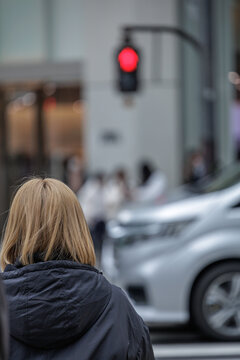 Candid urban street moment of blonde-haired women waiting for glowing red traffic light at busy crosswalk while car passes by in front of small group of people in bokeh background of gray winter day.