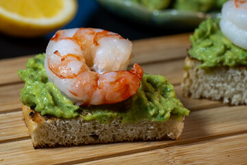 a single triangular toast bite topped with chunky mashed avocado and a succulent cooked shrimp, highlighting the texture of the bread and topping on a wooden board.