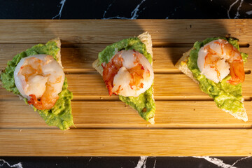three triangular toast appetizers topped with creamy mashed avocado and cooked shrimp, lined up neatly on a wooden slat serving board against a black