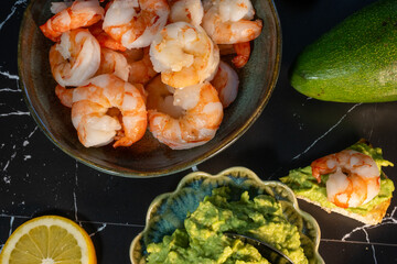 flat lay featuring a bowl of cooked shrimp, a decorative green bowl of mashed avocado, and a prepared toast bite, arranged on a black marble surface next to a fresh avocado and lemon slice.