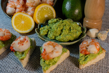 Triangular toast bites topped with mashed avocado and shrimp, arranged in front of a bowl of guacamole, lemon slices, and a pepper mill.