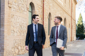 Two smiling young businessmen in suits walking and discussing work outside a modern office building