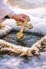 Close-up of a priest's hand dipping a golden ornate cross into an icy hole in the water; Epiphany celebration, Christian ritual of water blessing in winter with snow and frozen pine branches.
