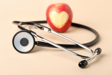 Ripe apple with carved heart and stethoscope on beige background, closeup