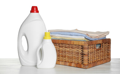 Laundry basket with clean clothes and bottles of detergents on light wooden table against white...