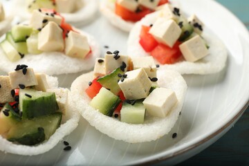 Tasty rice paper chips with vegetables, feta cheese and black sesame seeds on light blue table, closeup