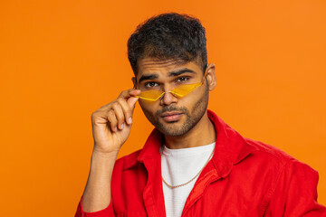 Proud Indian stylish man wearing sunglasses, charming smile, feels happy and successful. Portrait Arabian prideful young guy celebrating success victory, win, achievement isolated on orange background
