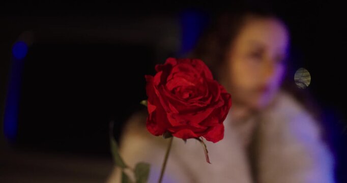 Close-up of a red rose sharply in focus while a woman appears blurred in the background, creating a cinematic romantic atmosphere. Concept for love, longing, breakup, and emotional storytelling.