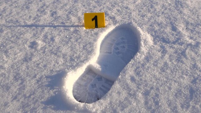 Slow cinematic orbiting around a boot print or footprint in fluffy white winter snow with a yellow Crime scene investigation marker next to it, sunny day.