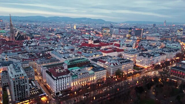 Aerial landscape static drone shot of Vienna cityscape at night in winter, showing illuminated buildings, glowing streets, flowing traffic, and city lights reflecting across rooftops under winter sky