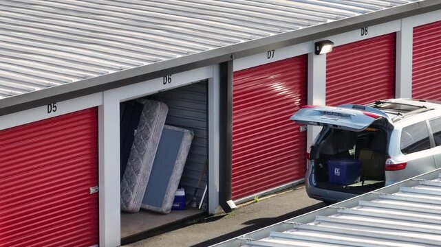 Aerial view of a self storage facility in United States, showing red roll-up garage units, an open storage space with mattresses inside. Car loaded at rental unit.