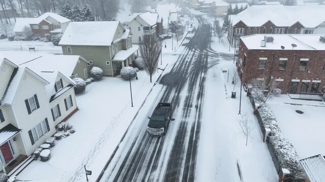 Black pickup truck on snowy road in American neighborhood. Aerial tilt down shot. Snowfall in quiet residential area of town. Winter season in USA.